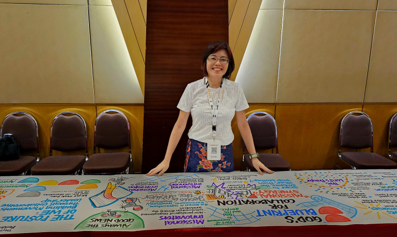 Wendy, a visual scribe from Singapore, stands in front of her illustrated scroll capturing key moments from the Arise Leadership Summit 2025 in Indonesia, where she served by documenting the four-day event through live visual storytelling.