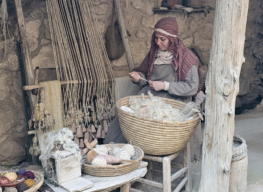A woman portrayed in first-century dress works wool by hand at Nazareth Village, demonstrating textile skills used to produce clothing and household fabrics in Galilee during the time of Jesus.