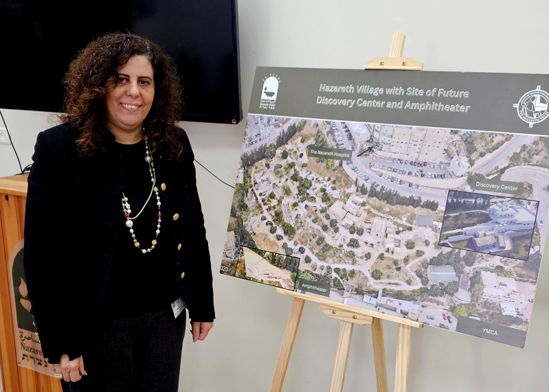Maha Sayegh, director of Nazareth Village, stands beside a visual showing an aerial view of the site with a rendering of the planned visitor center and amphitheater as part of the village’s expansion plans.