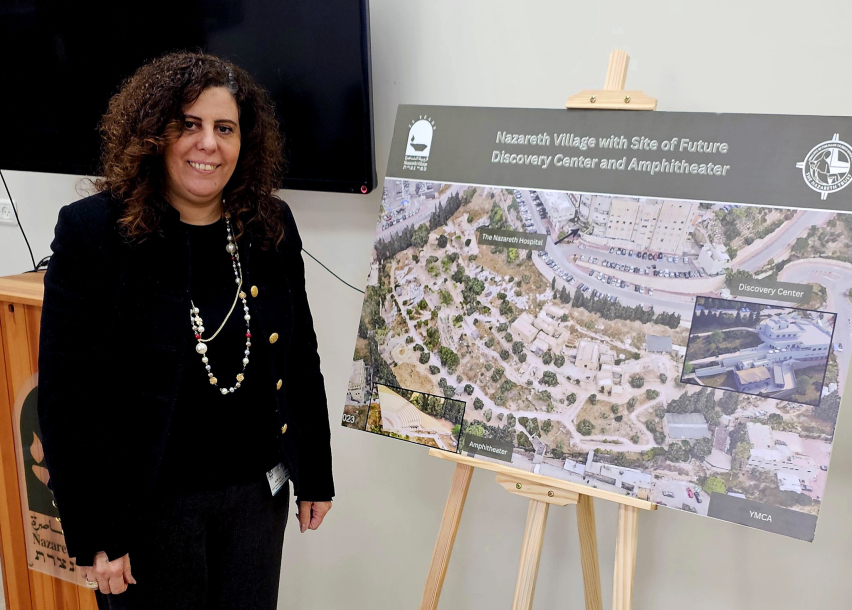 Maha Sayegh, director of Nazareth Village, stands beside a visual showing an aerial view of the site with a rendering of the planned visitor center and amphitheater as part of the village’s expansion plans.