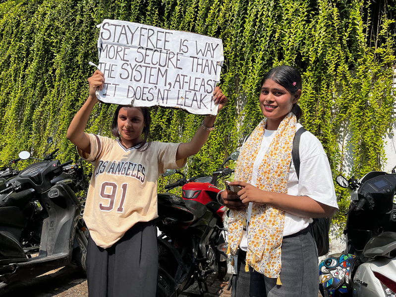 Students protest Indias examination system in Delhi, India on July 23, 2026. One demonstrator holds a sign reading Stayfree is way more secure than the system, at least it doesnt leak — a reference to the menstrual pad brand and a pointed jab at the e