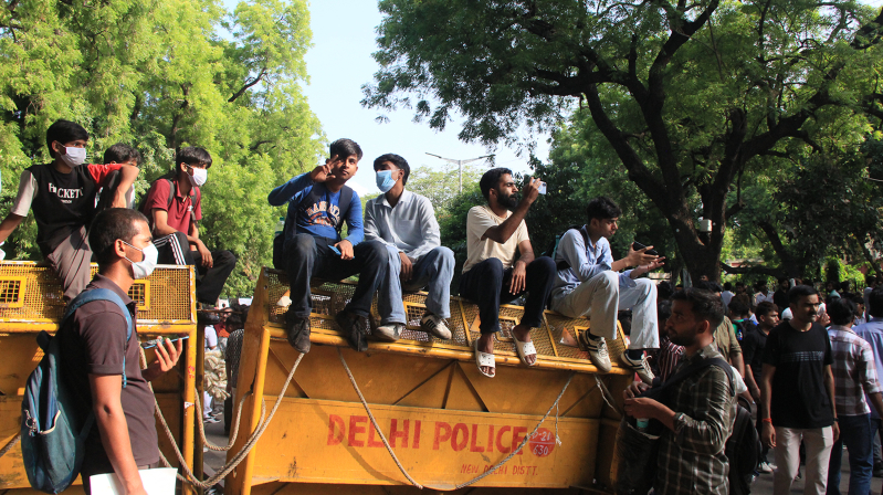 Demonstrators climb atop a Delhi Police barricade to watch proceedings at Jantar Mantar in New Delhi during protests led by the youth-led Cockroach Janta Party.