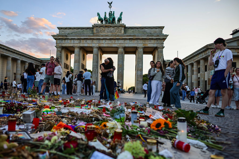 Tourists and visitors observe and pay respects at a makeshift memorial set in Pariser Platz in front of the Brandenburg gate, one week after the Christopher Street Day pride parade attack on August 2, 2026 in Berlin, Germany.